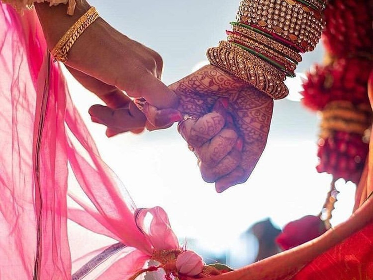 The bride and groom's hands clasped, her mehndi and bangles catching the light, their dupattas knotted below
